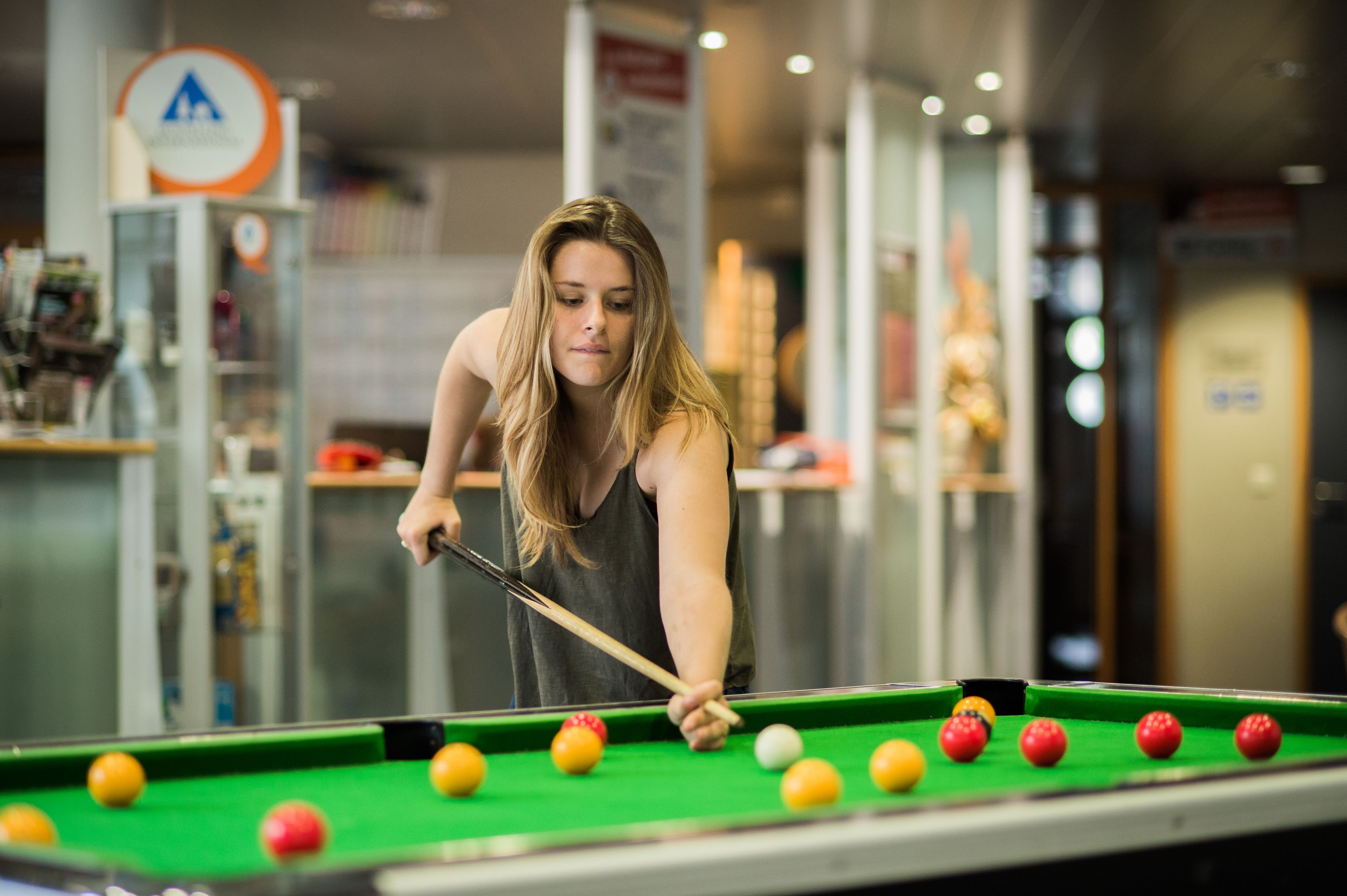 A person playing pool in a room with a green pool table, shelves with various items, and a sign with a triangle symbol.