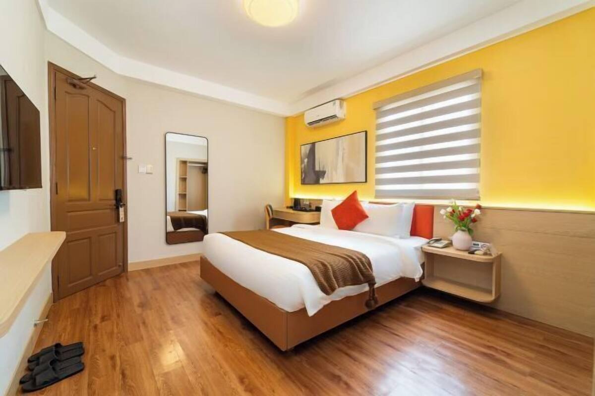 Ein Hotelzimmer mit Holzboden, einem Bett mit weißer Bettwäsche, einem Nachttisch mit Blumen in einer Vase, einem Spiegel und einem Fenster mit Jalousien.