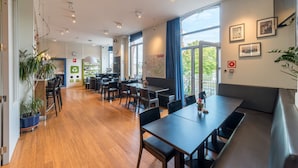 A modern dining area with wooden flooring, black tables, and chairs. There are potted plants and a large window with a view of greenery. - Auberge de Jeunesse de Namur - Hostel (Namur)