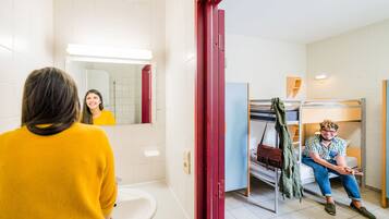 Baño con inodoro, lavabo y espejo. Se observa una cama litera con una chaqueta verde colgada.