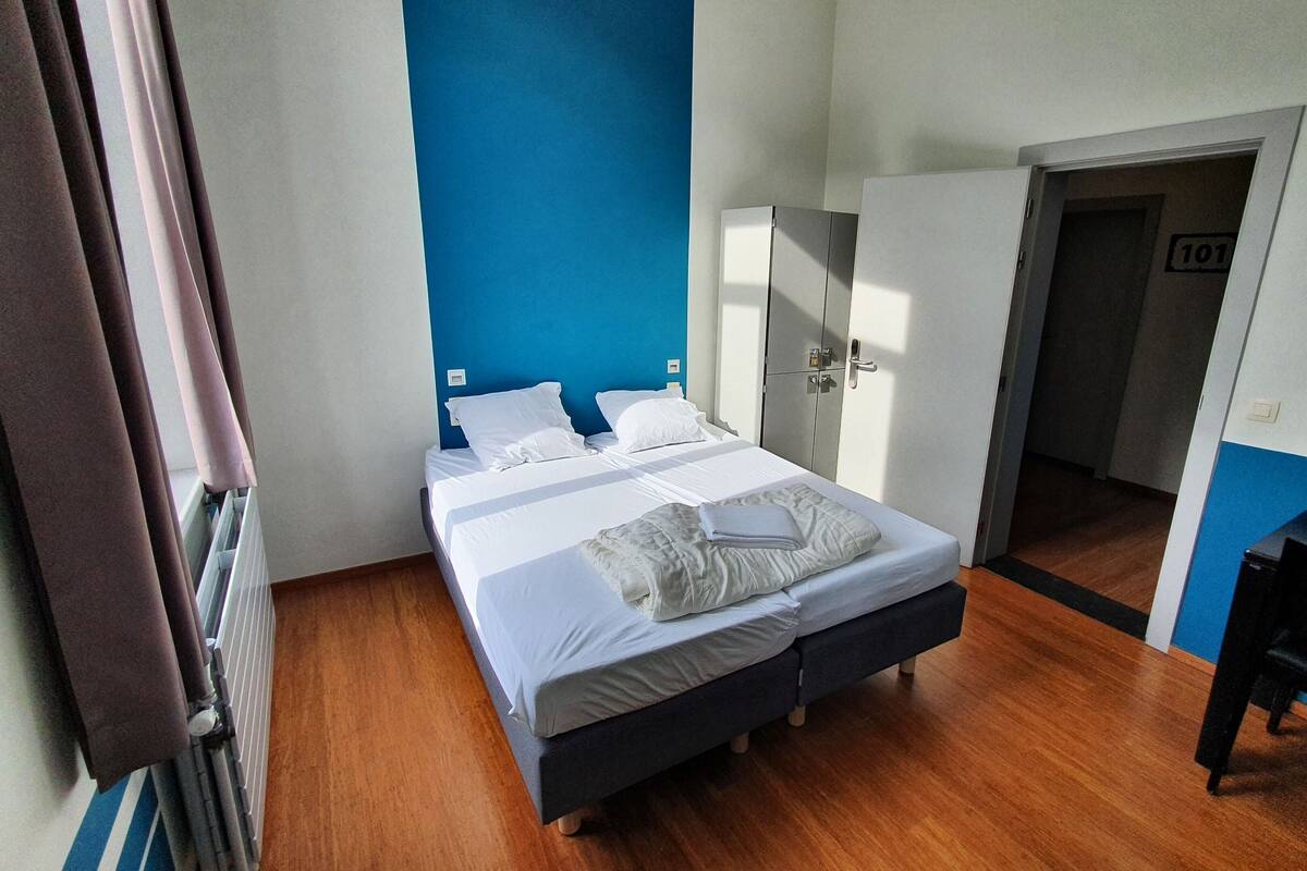 Un lit double avec du linge de lit blanc, un mur d’accent bleu, une armoire et un bureau.