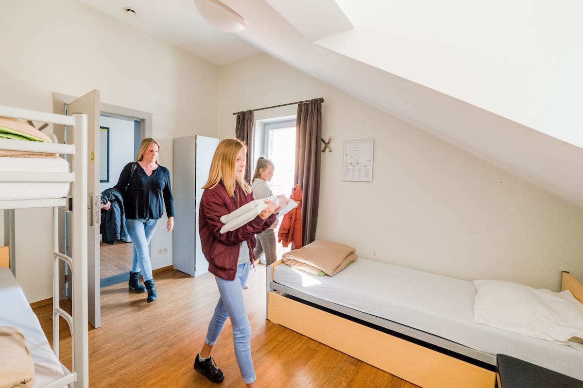 Une chambre de dortoir avec des lits superposés, un lit simple, un plancher en bois, une fenêtre avec des rideaux et un luminaire fixé au mur.