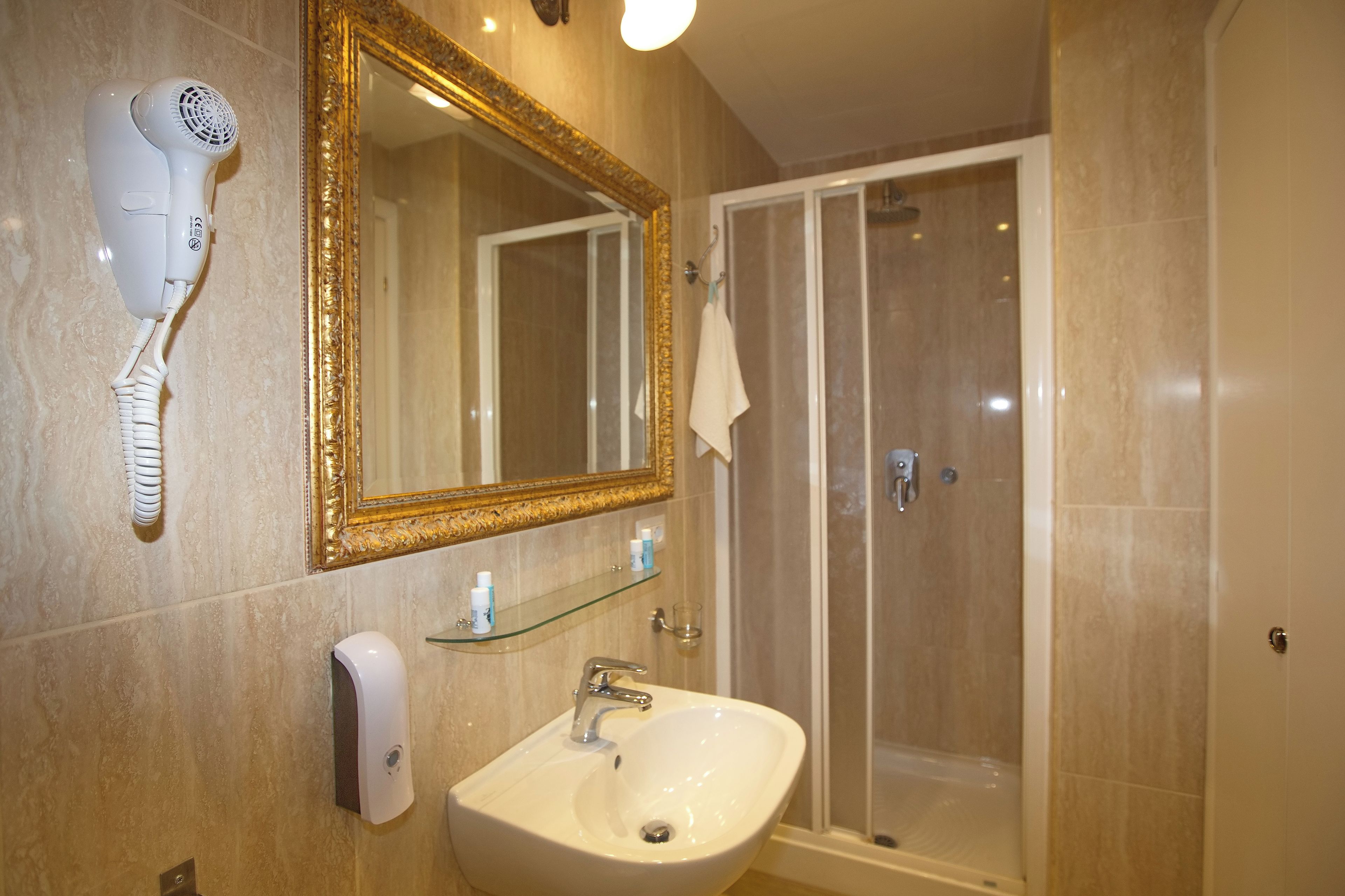 Een badkamer met een witte wastafel, een spiegel met gouden lijst, een haardroger en een douchecabine van glas.