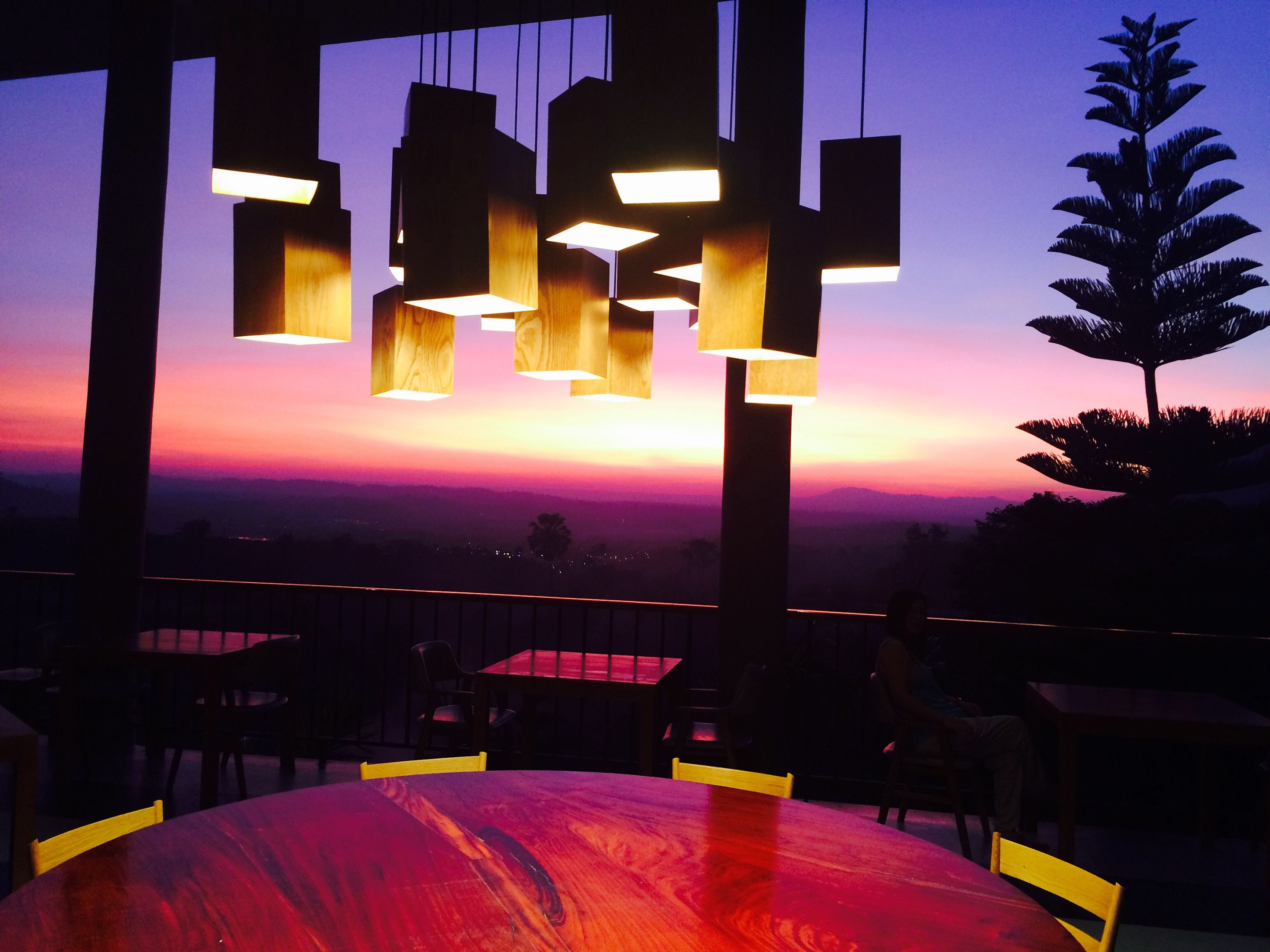 Un restaurante con vista al atardecer, con mesas y sillas de madera, y lámparas colgantes.