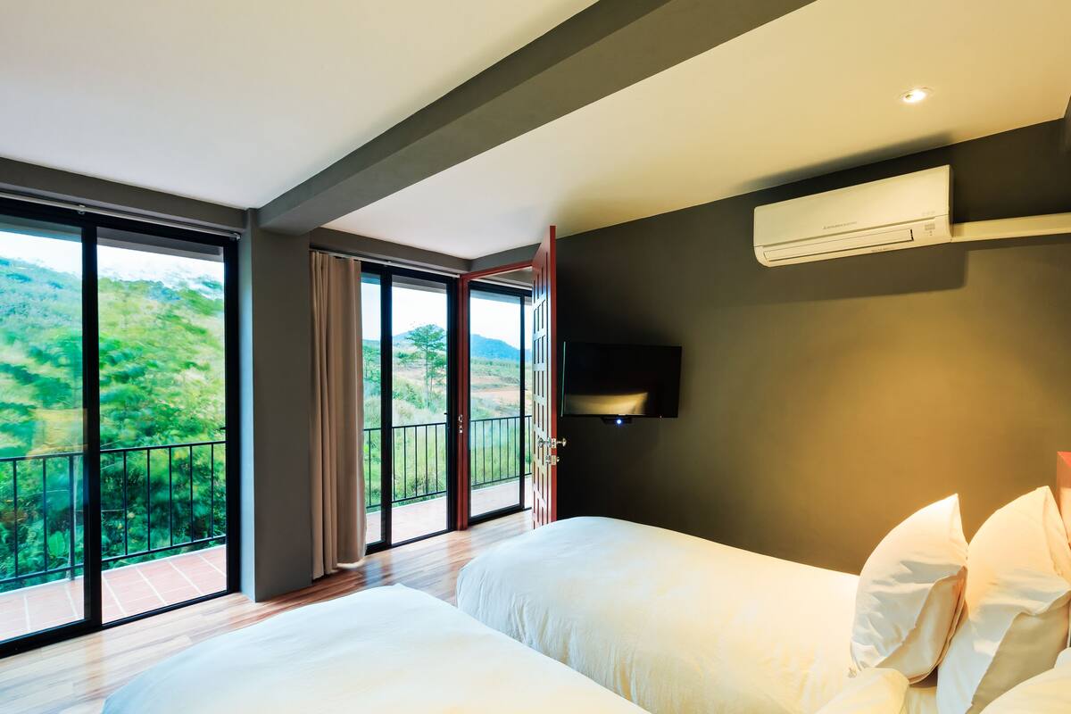 A hotel room with two beds, a large window with a view of greenery, and an air conditioning unit on the wall.