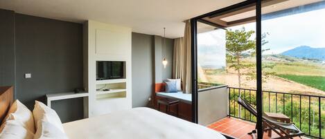 A bedroom with a large bed, a TV, a desk, and a balcony with a view of the countryside.
