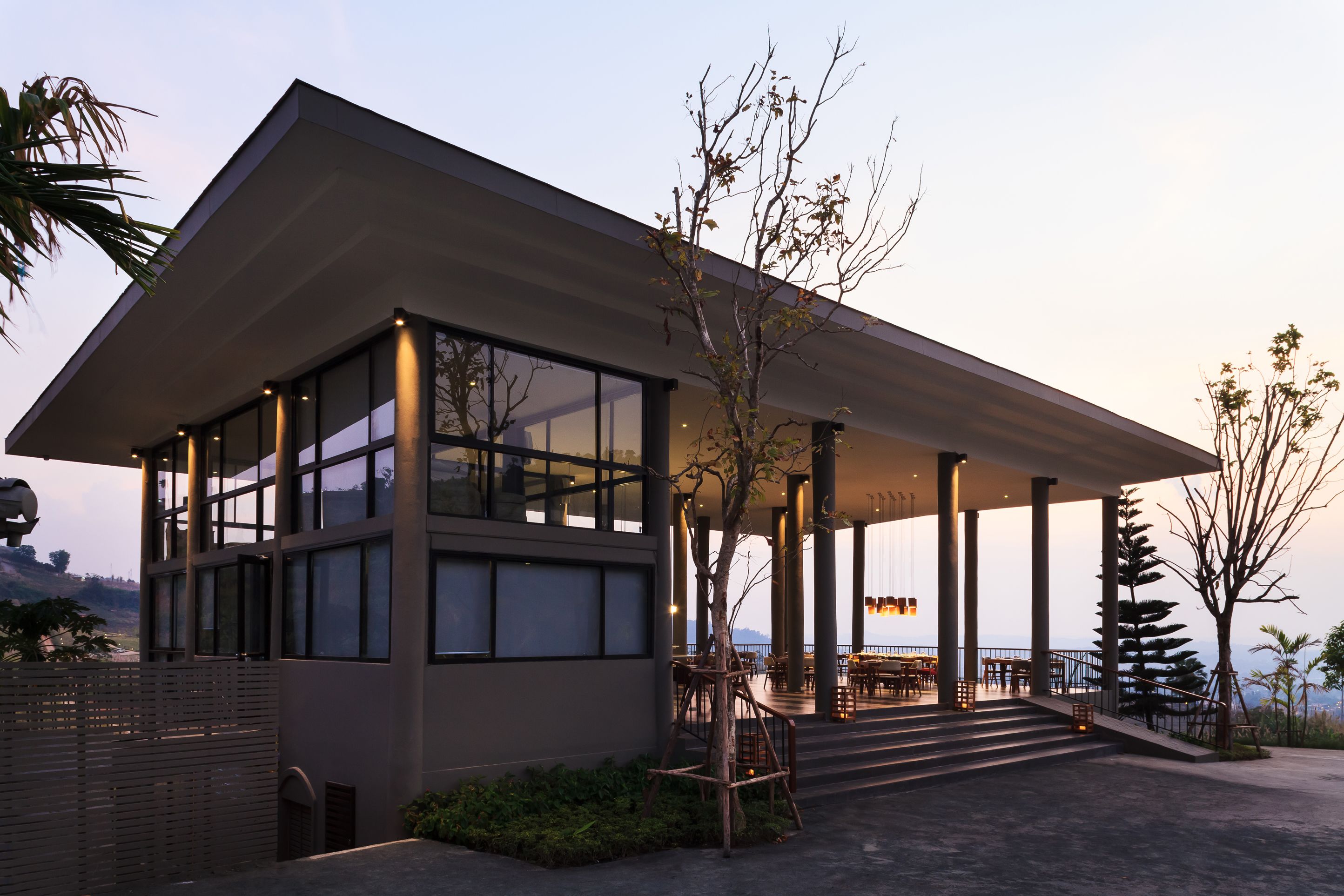 A modern building with large windows, a covered outdoor area with seating, and a view of the landscape.