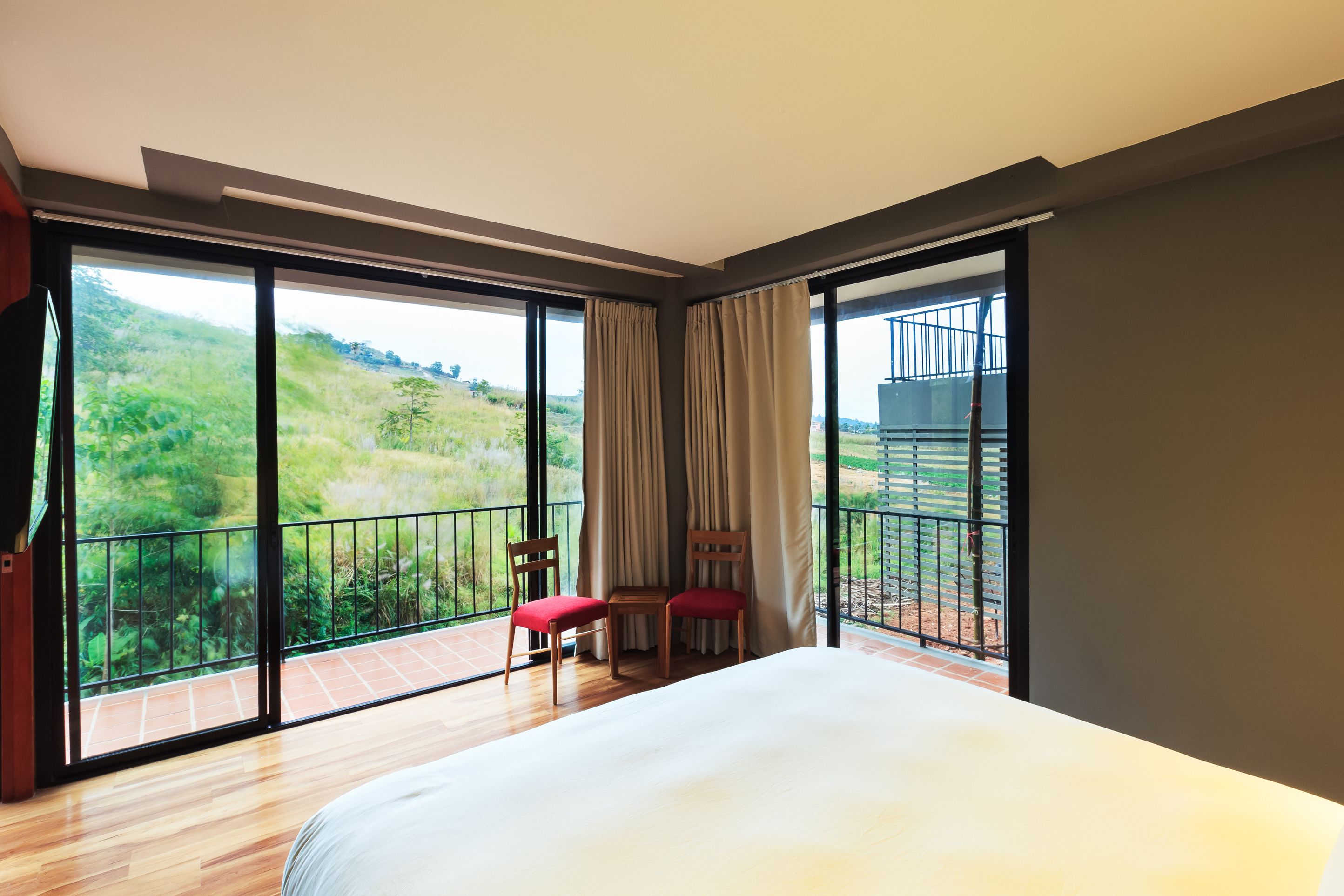 A bedroom with a large bed, two red chairs, and a balcony with a view of greenery.