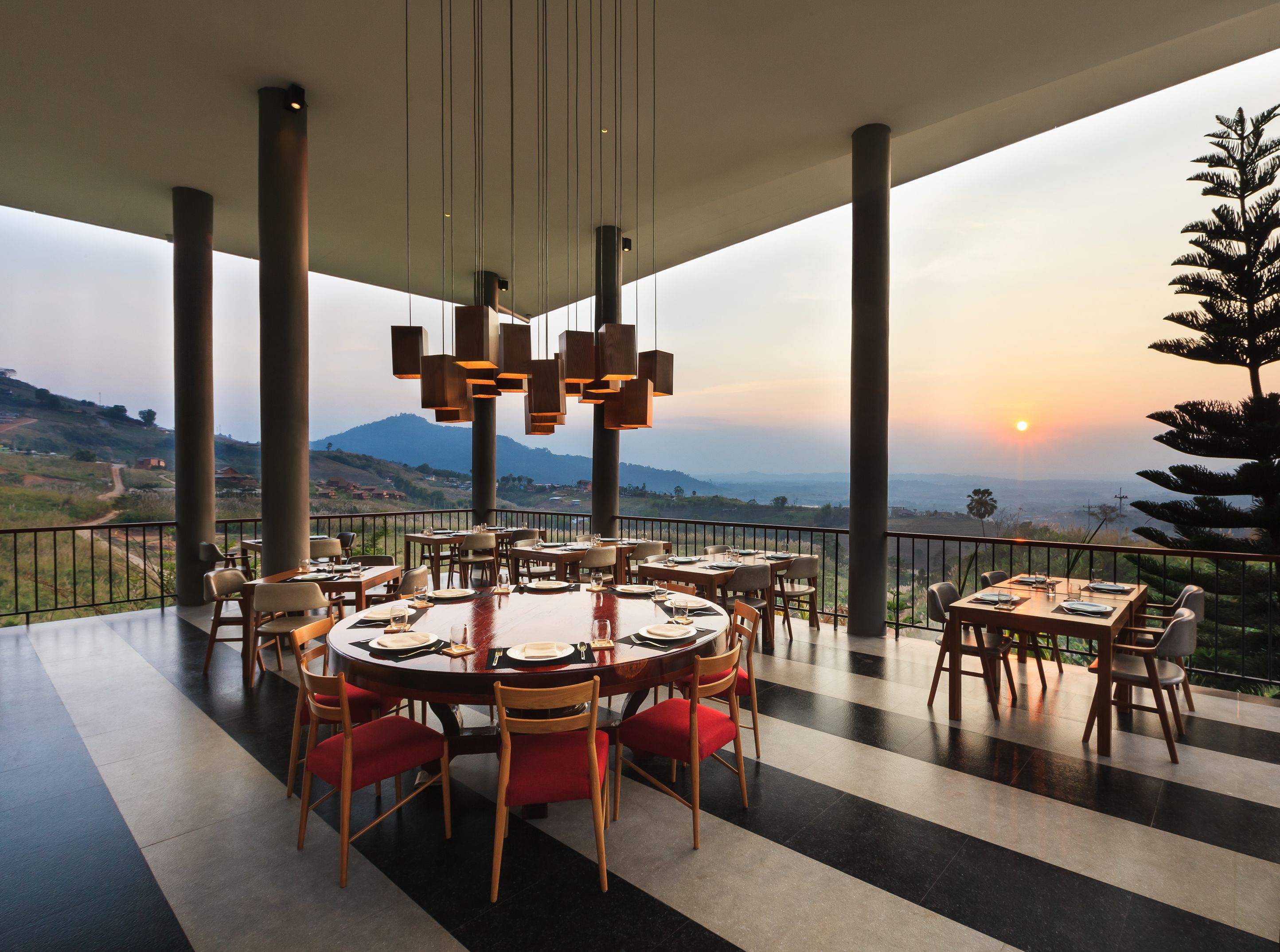 A restaurant with round tables and red chairs, large pillars, and a view of mountains and a sunset.