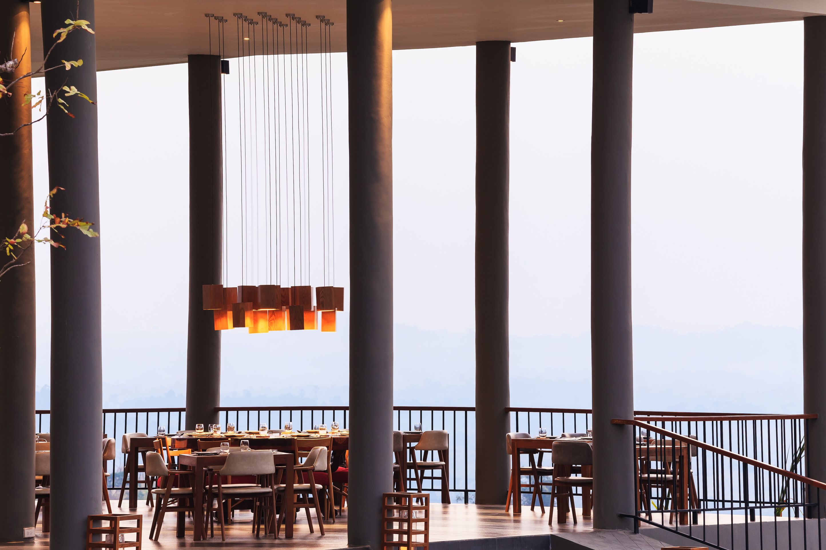 A restaurant with high ceilings, large pillars, and hanging lights. Tables and chairs are arranged for dining.