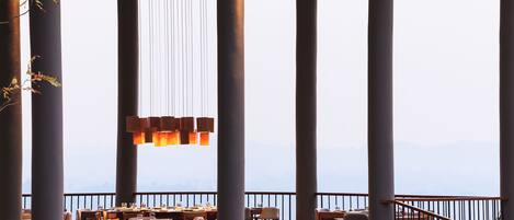 A restaurant with high ceilings, large pillars, and hanging lights. Tables and chairs are arranged for dining.