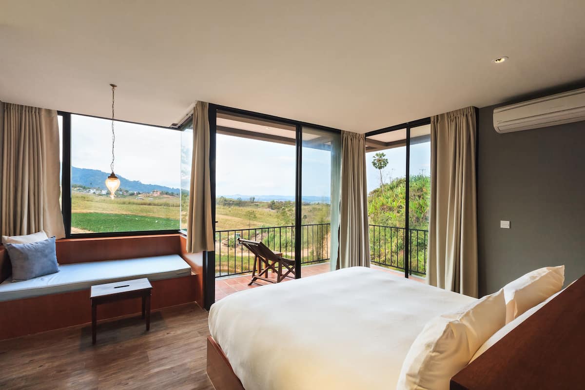 A bedroom with a large bed, a bench, and a view of a green landscape through large windows.