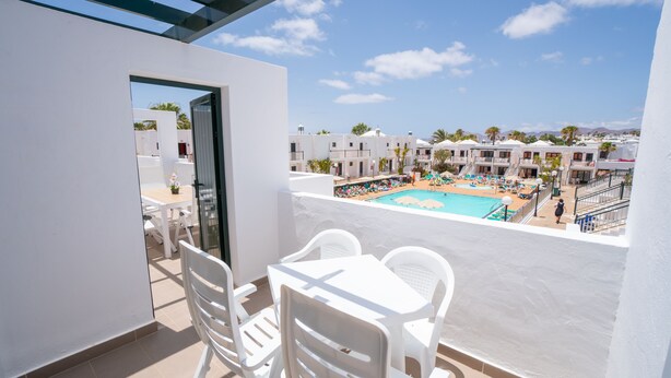 Un balcón con sillas y una mesa blancos, con vistas a una piscina con tumbonas y sombrillas, rodeado de edificios blancos y palmeras.