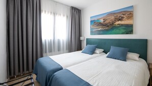 Una habitación de hotel con dos camas, un ventanal con cortinas, un cuadro de un paisaje costero y un cabecero.