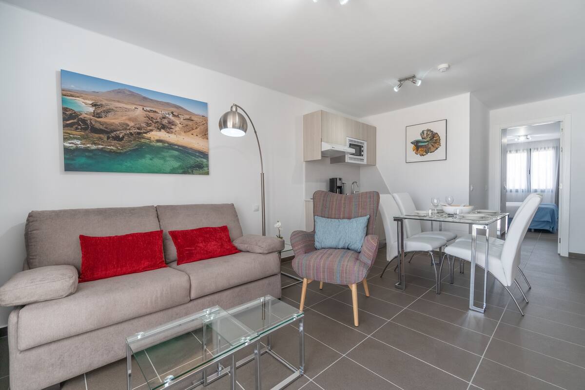 Ein modernes Wohnzimmer mit einer Couch, einem Sessel und einem Glastisch. Es gibt einen Essbereich mit Tisch und Stühlen. Ein Landschaftsbild hängt an der Wand.