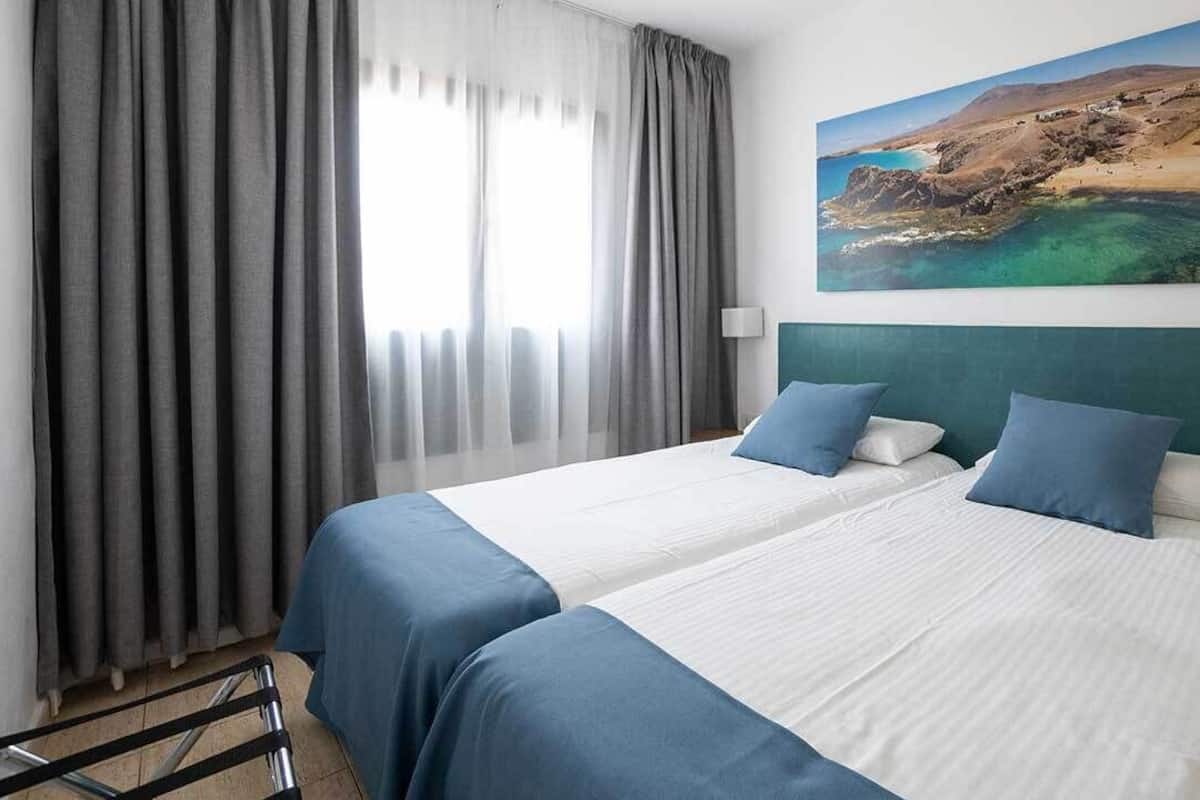 A hotel room with two beds, grey curtains, a painting of a coastal landscape, and a green headboard.