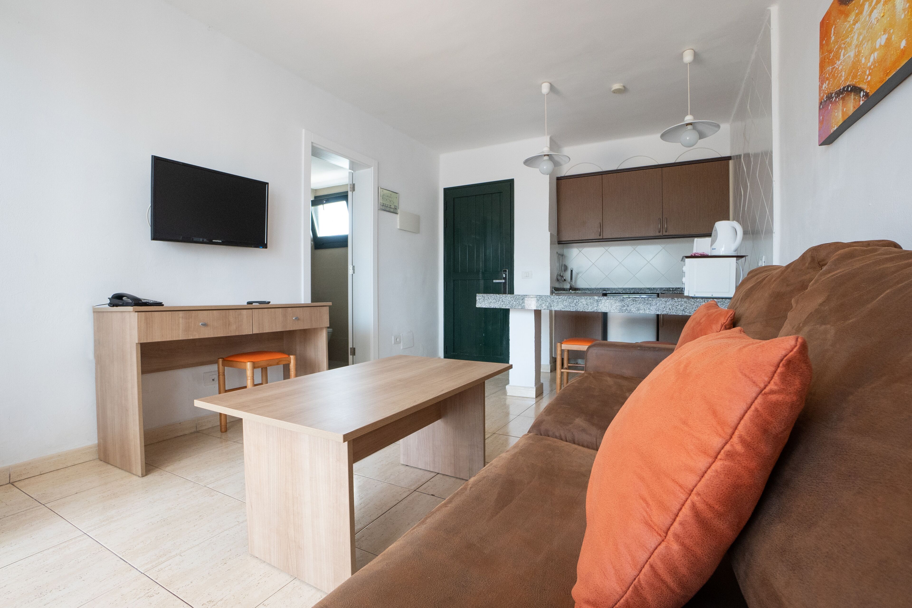 Een moderne keuken met een eethoek, een aan de muur bevestigde televisie en een bank met oranje kussens.