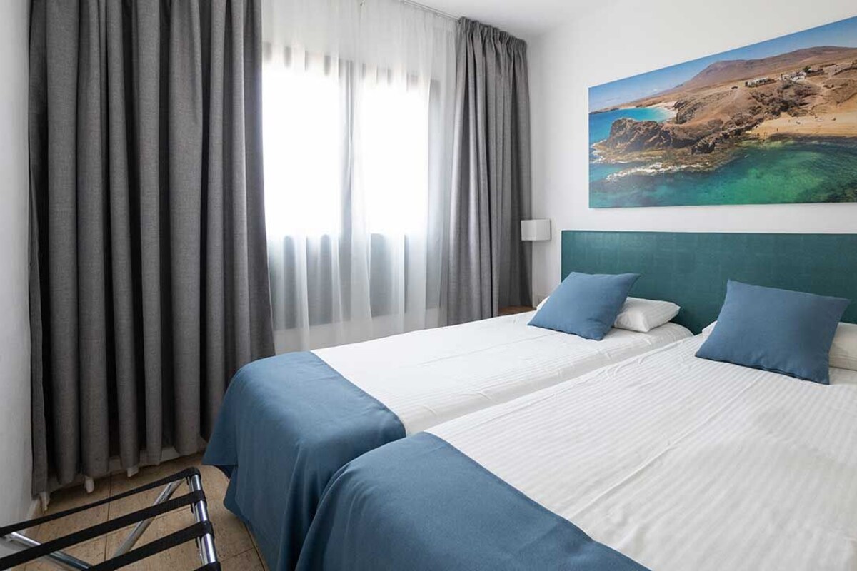 A hotel room with two beds, grey curtains, a painting of a coastal landscape, and a green headboard.