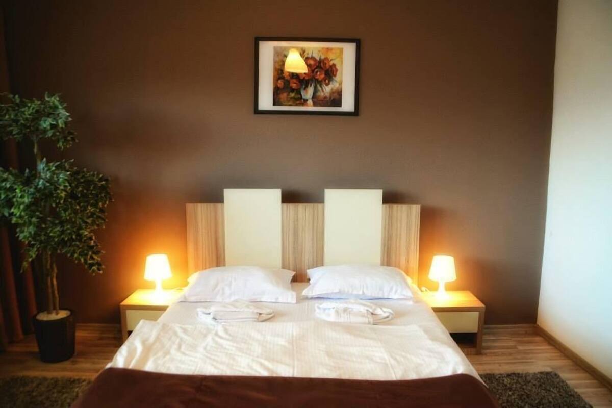 A neatly made double bed with white linens, two bedside lamps, a framed painting above the bed, and a potted plant to the left.