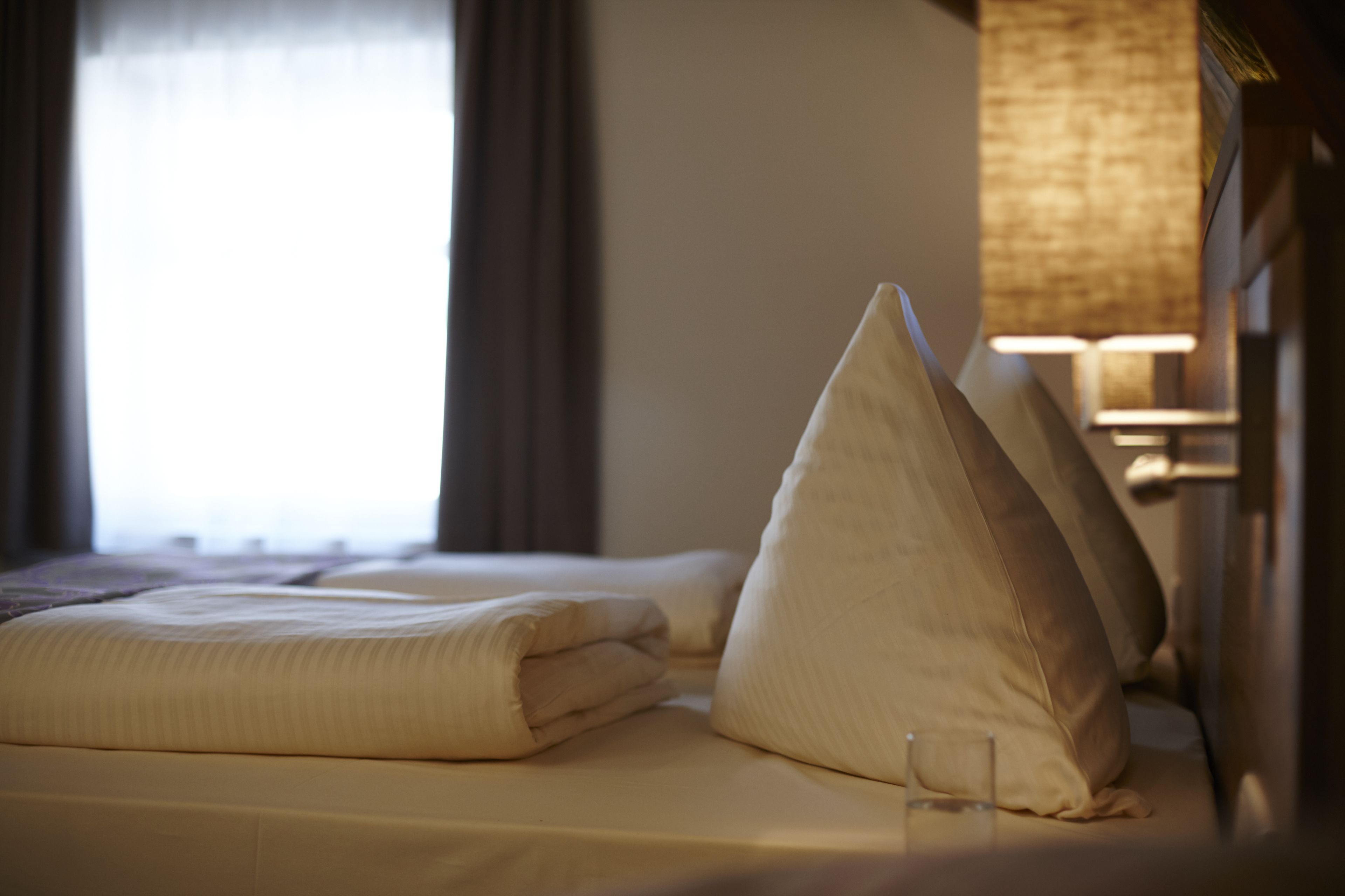 A neatly made bed with pillows and a folded blanket, a lamp, and a window with curtains.