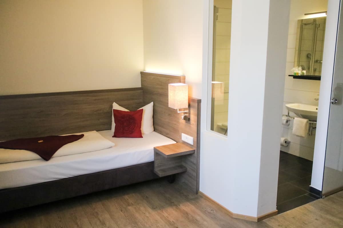 Une chambre d’hôtel moderne, équipée d’un lit, d’une table de chevet, d’une lampe et offrant une vue sur la salle de bain.
