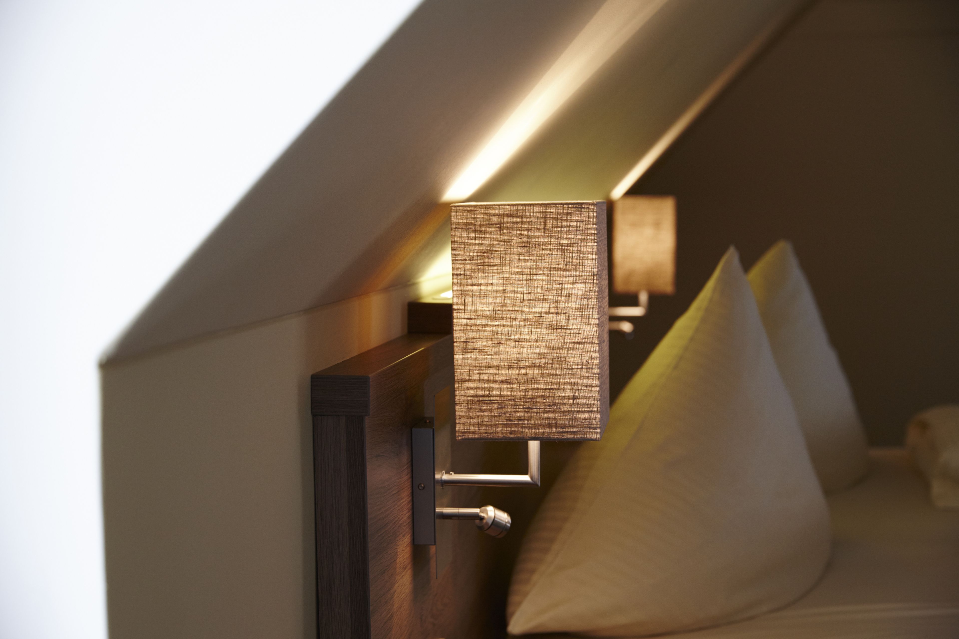 A modern hotel room with a sloped ceiling, a wall-mounted lamp, and a bed with pillows.