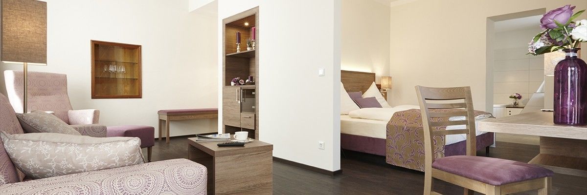 A modern hotel room with a sofa, a dining table, a bed, and a small kitchen area.