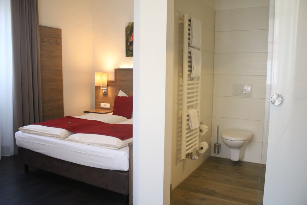 Una habitación de hotel con cama, mesita de noche, lámpara y vistas al baño.