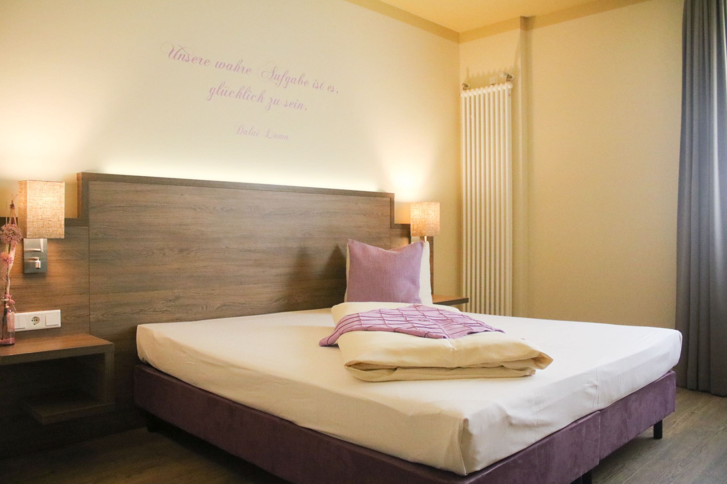 Una cama bien tendida en una habitación de hotel, con cabecera de madera, una lámpara de pared y un cojín decorativo.