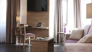 A modern hotel room with a wooden desk, a comfortable sofa, a flat-screen TV mounted on the wall, and a window with curtains.