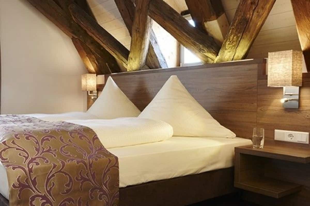 Une chambre d’hôtel avec un lit, une chaise, un bureau, une lampe et une fenêtre avec des rideaux.