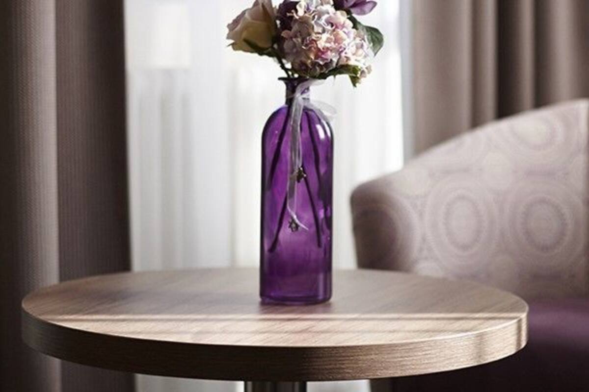 Une table ronde en bois, sur laquelle est posé un vase violet rempli de fleurs, se trouve à côté d’un fauteuil à motifs.