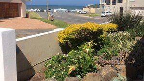 Una calle residencial con casas, vista despejada al océano y un jardín cuidado con rocas y plantas.