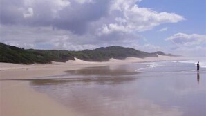 Una playa con una extensa orilla arenosa, olas suaves y una persona de pie a la derecha.