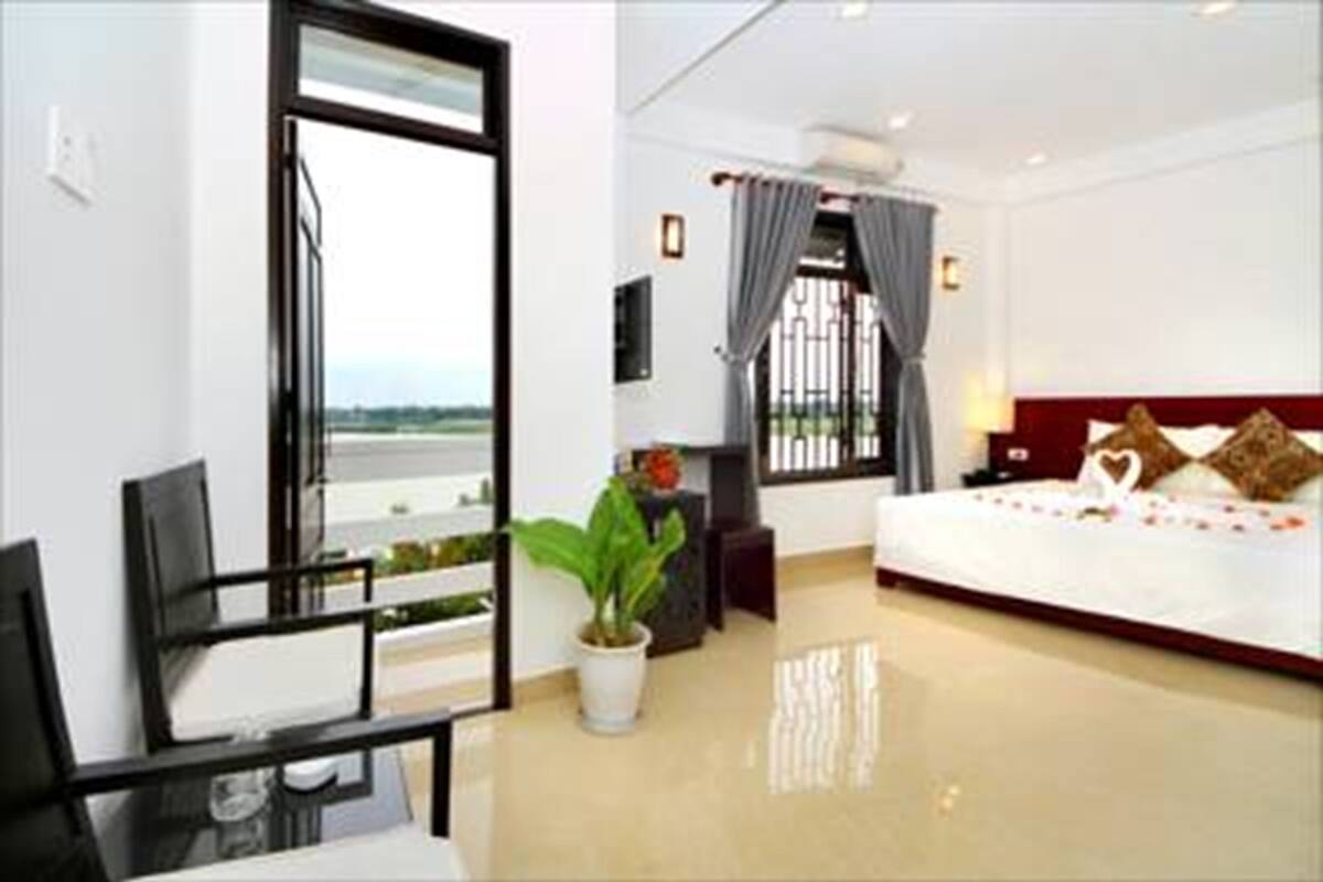 A hotel room with a bed, a desk, a chair, a potted plant, and a view of the outdoors.