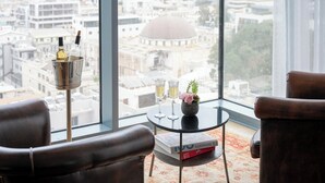 A modern room with leather chairs, a small table with a book, two champagne flutes, and a decorative vase. A large window offers a view of a cityscape and a dome structure. - Hotel Rothschild 22 - FATTAL COLORS (Tel Aviv)