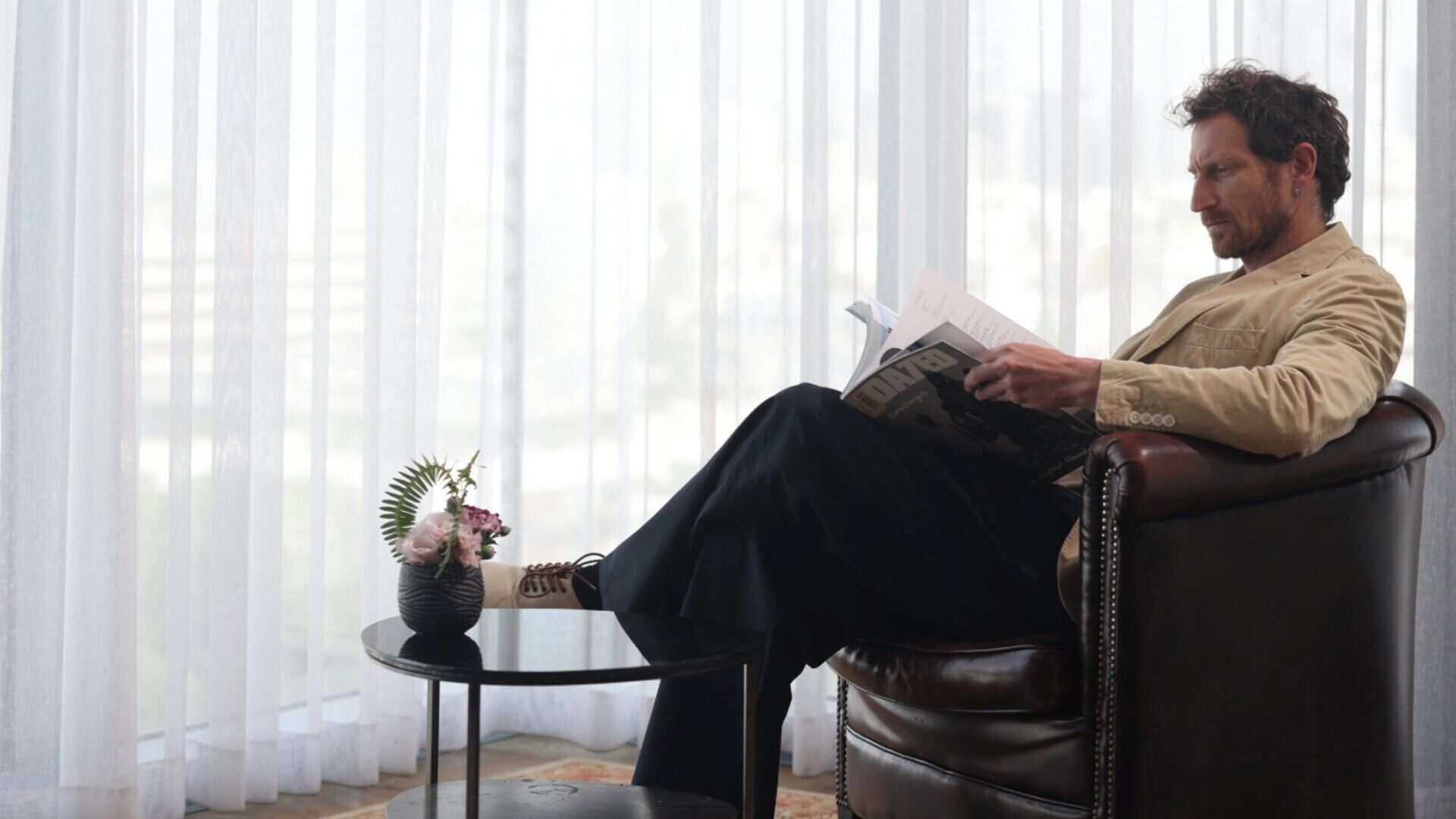 Un hombre sentado en un sillón de cuero leyendo una revista en una habitación con cortinas translúcidas y una mesita con un jarrón de flores.