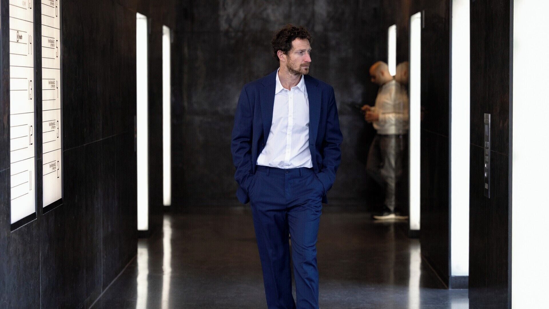 A man in a blue suit walking down a hallway with a dark floor and walls, and a person in the background.