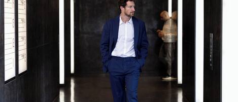 A man in a blue suit walking down a hallway with a dark floor and walls, and a person in the background.