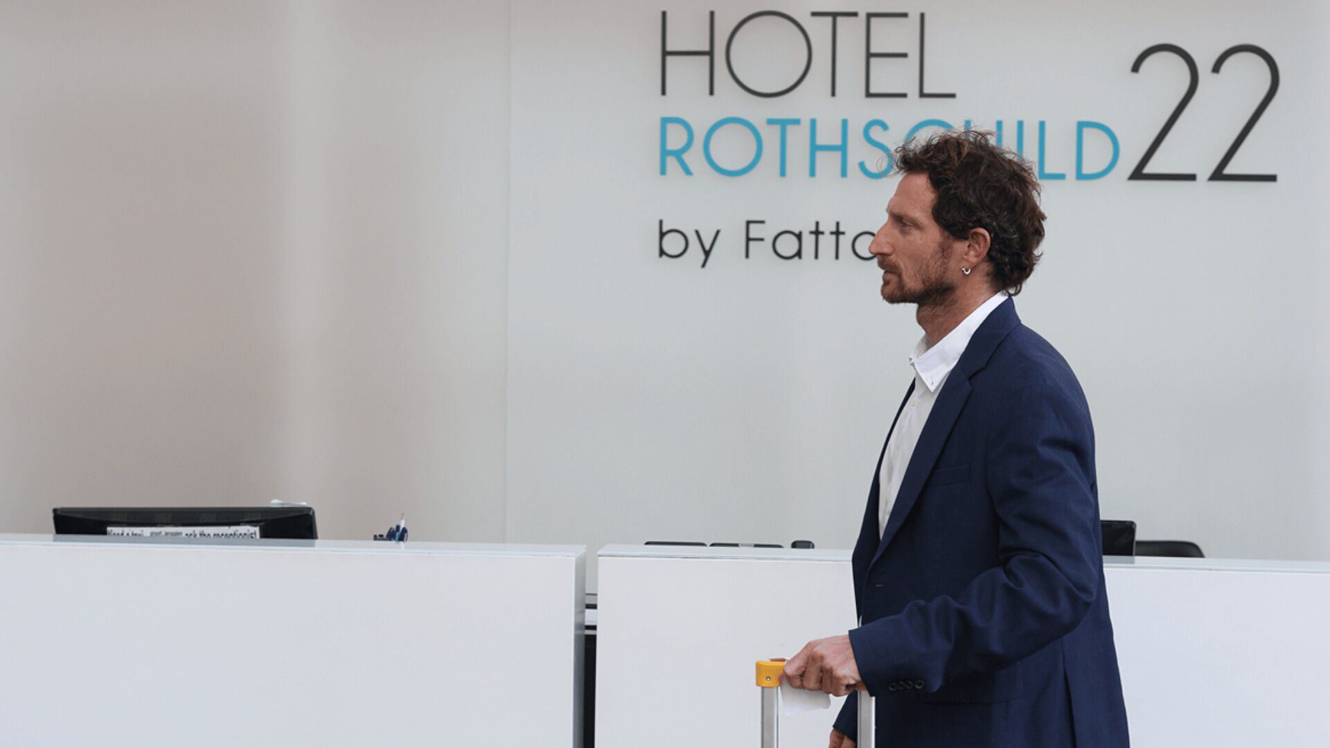 A man in a blue suit standing at a podium with a sign reading "HOTEL ROTHSCHILD 22 by Fattori" in the background.