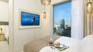 Quarto de hotel moderno com uma janela grande que oferece vista para a cidade, uma cama bem arrumada, uma mesa de cabeceira com uma garrafa e copos, e um quadro pendurado na parede.