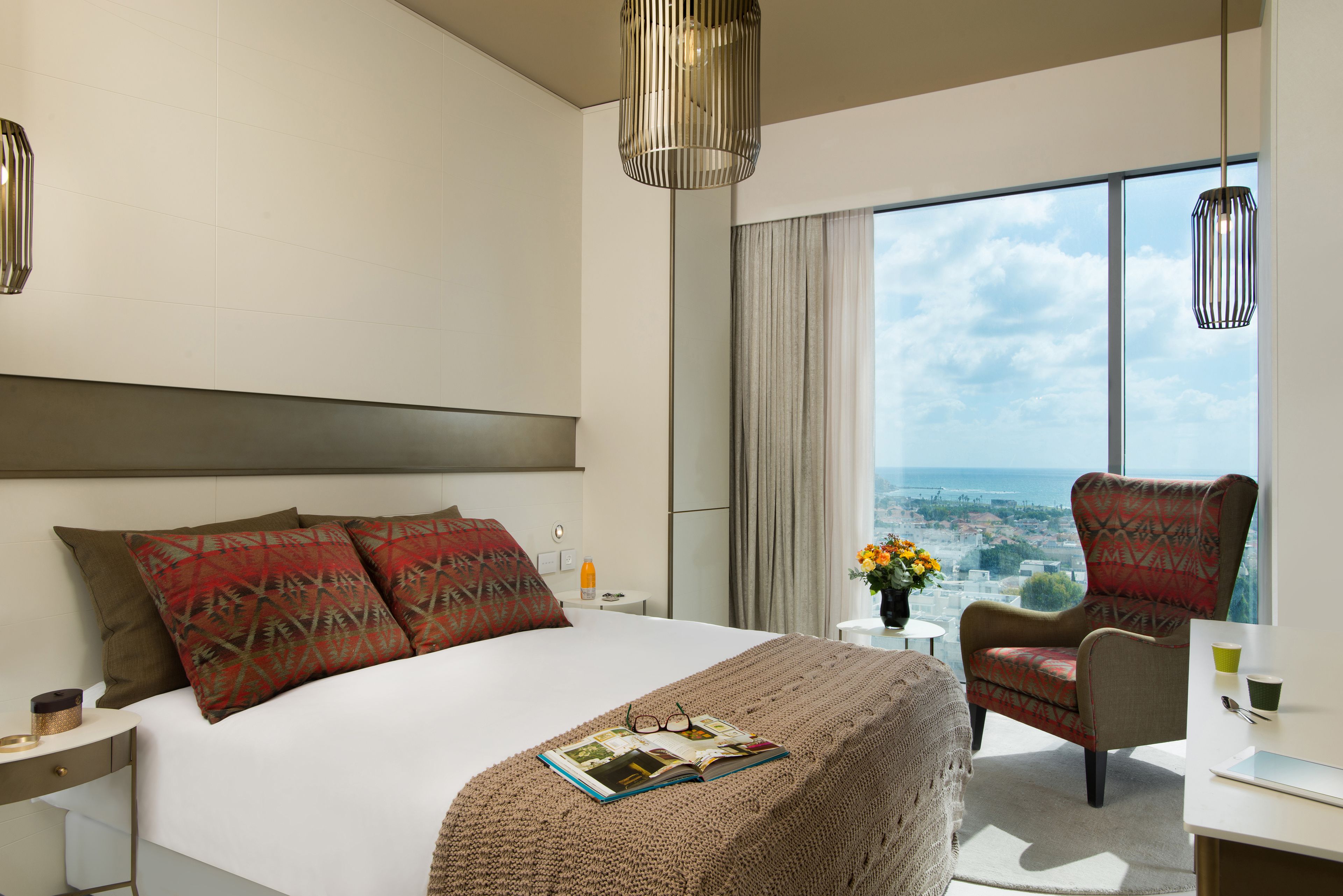 Quarto de hotel moderno com uma cama grande, uma poltrona, uma escrivaninha e vista para a cidade.