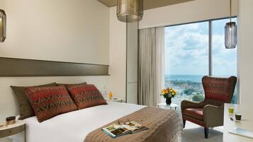 Quarto de hotel moderno com uma cama grande, uma poltrona, uma escrivaninha e vista para a cidade.