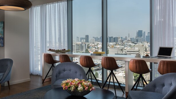 A modern dining area with a city view, a laptop on the table, and a bowl of flowers. - Hotel Rothschild 22 - FATTAL COLORS (Tel Aviv)