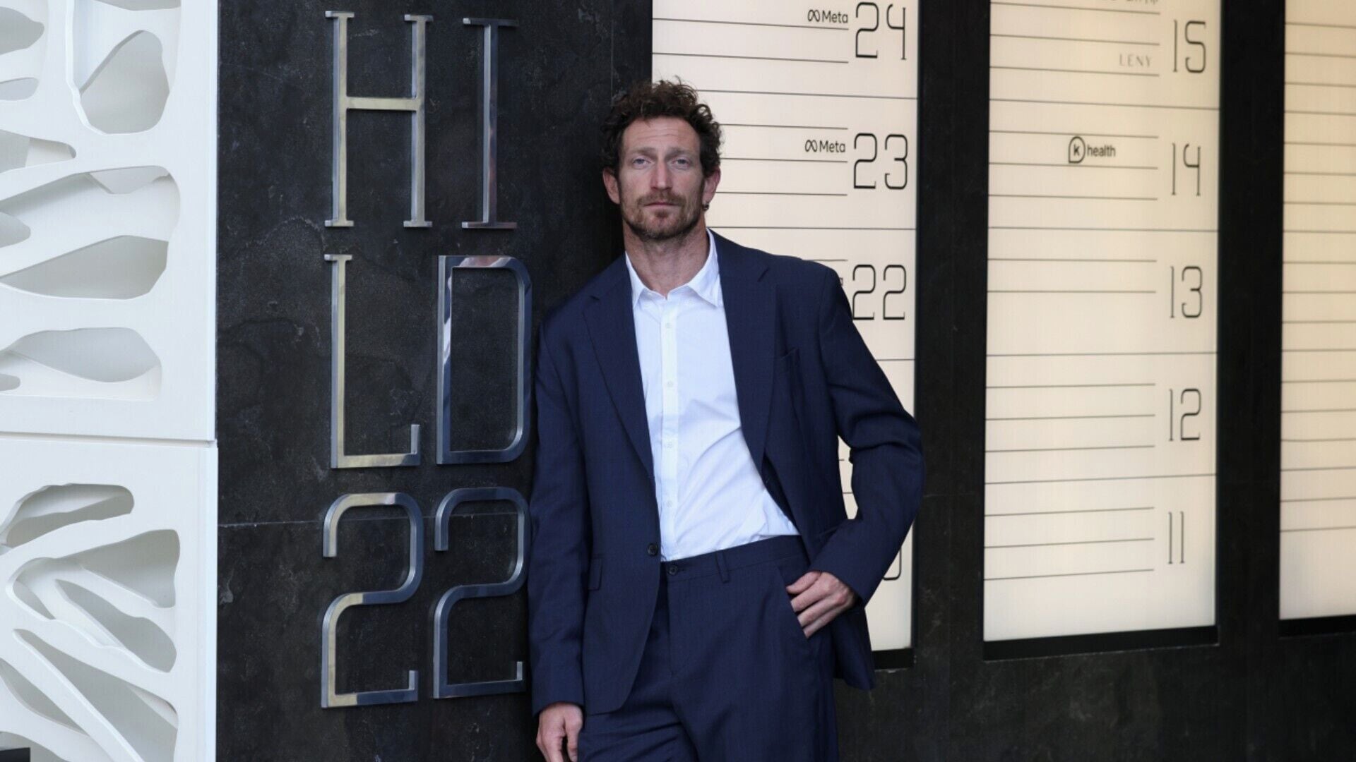 A man in a suit standing in front of a building with a large number '22' and the letters 'HI LD' visible.