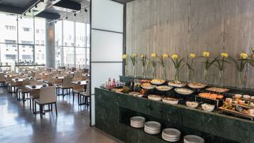 Un vaste espace dédié à la salle à manger, avec de nombreuses tables et chaises, un buffet proposant une variété de plats et de grandes fenêtres laissant entrer la lumière naturelle.
