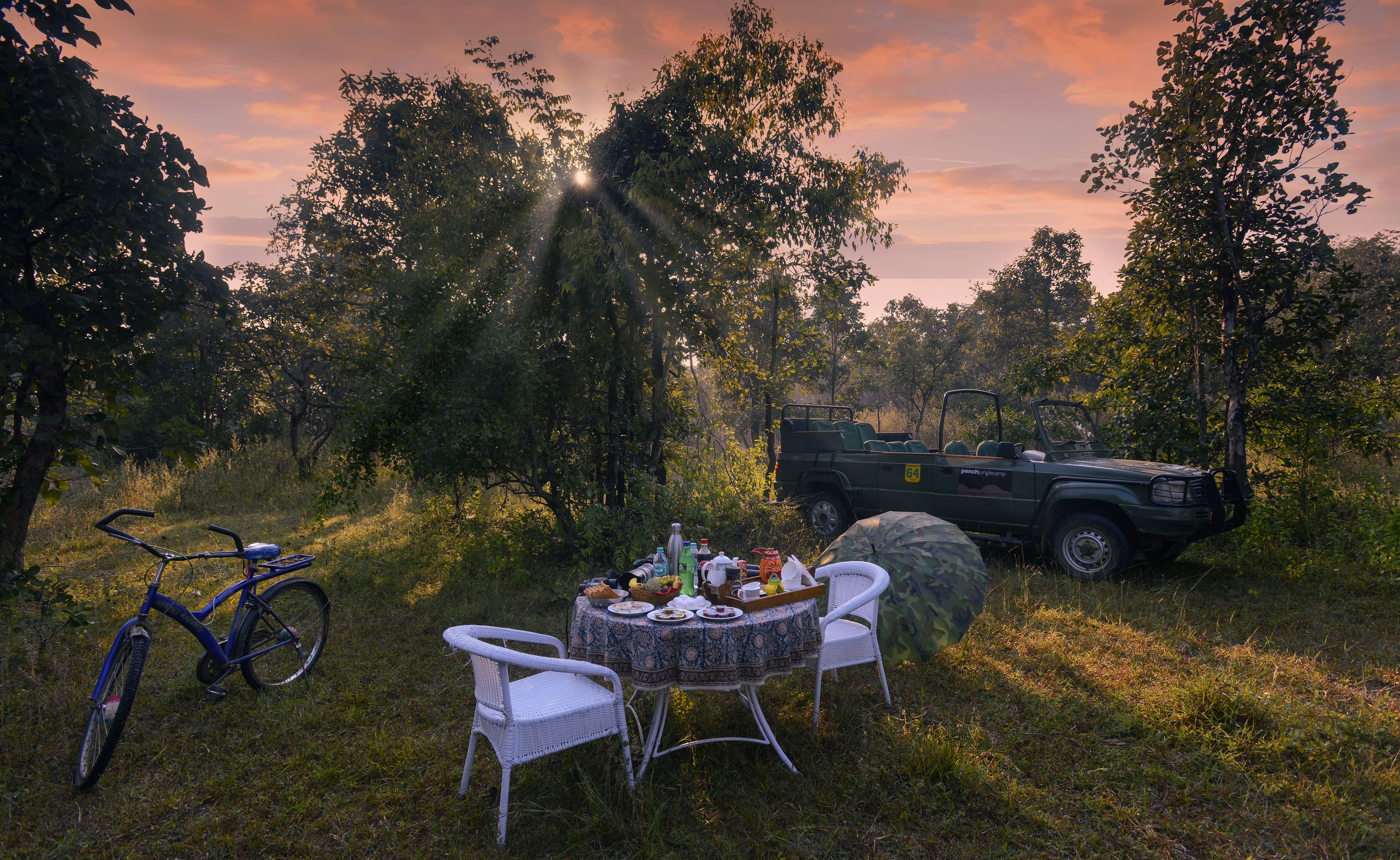 A table set outdoors with chairs, a bicycle, and a vehicle in a natural setting.