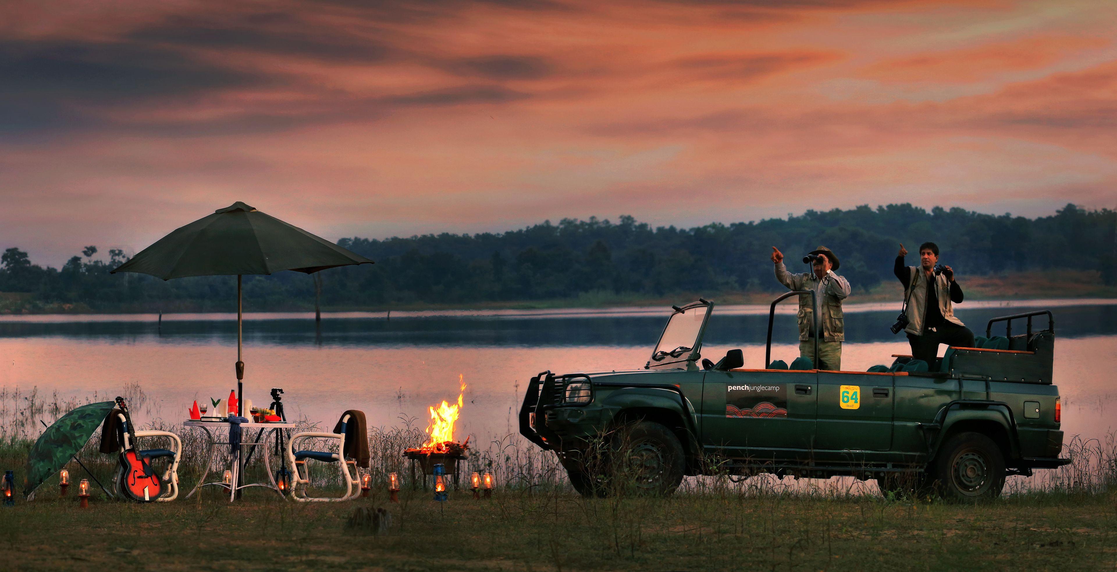 A green vehicle with a camper and a table with a fire in the foreground, a person standing on the vehicle, and a lake in the background.