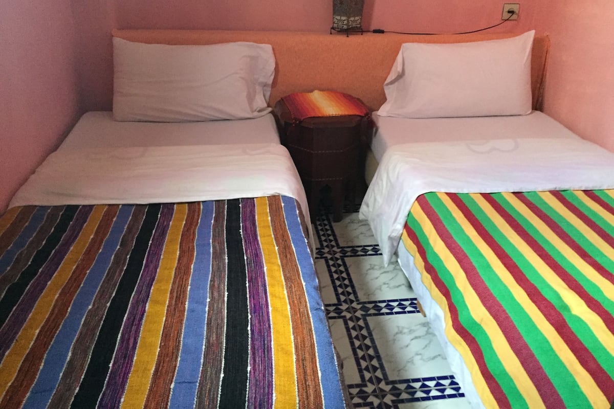A room with two beds, each with a different striped bedspread. There is a small table between the beds, and a wall-mounted shelf above the table.