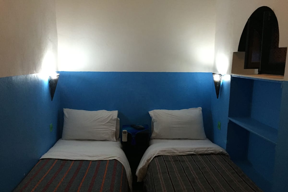 A small, narrow room with two single beds, each with a pillow and a striped blanket. There are two bedside tables with lamps, and the walls are painted blue.